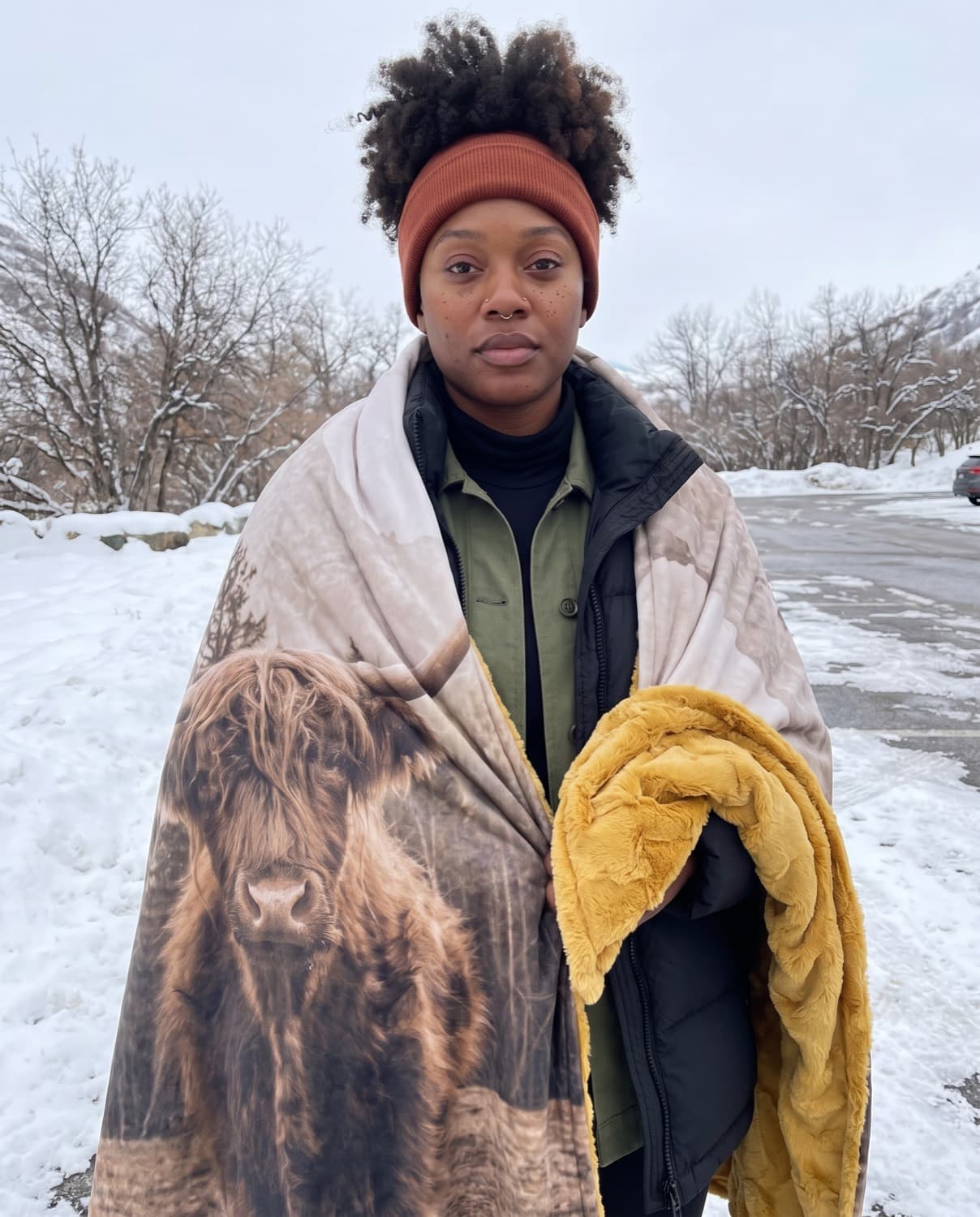 A woman in a rust headband stands in a snowy Utah canyon wrapped in the Highland Peak blanket, the cow printed across it and the golden plush over her arm