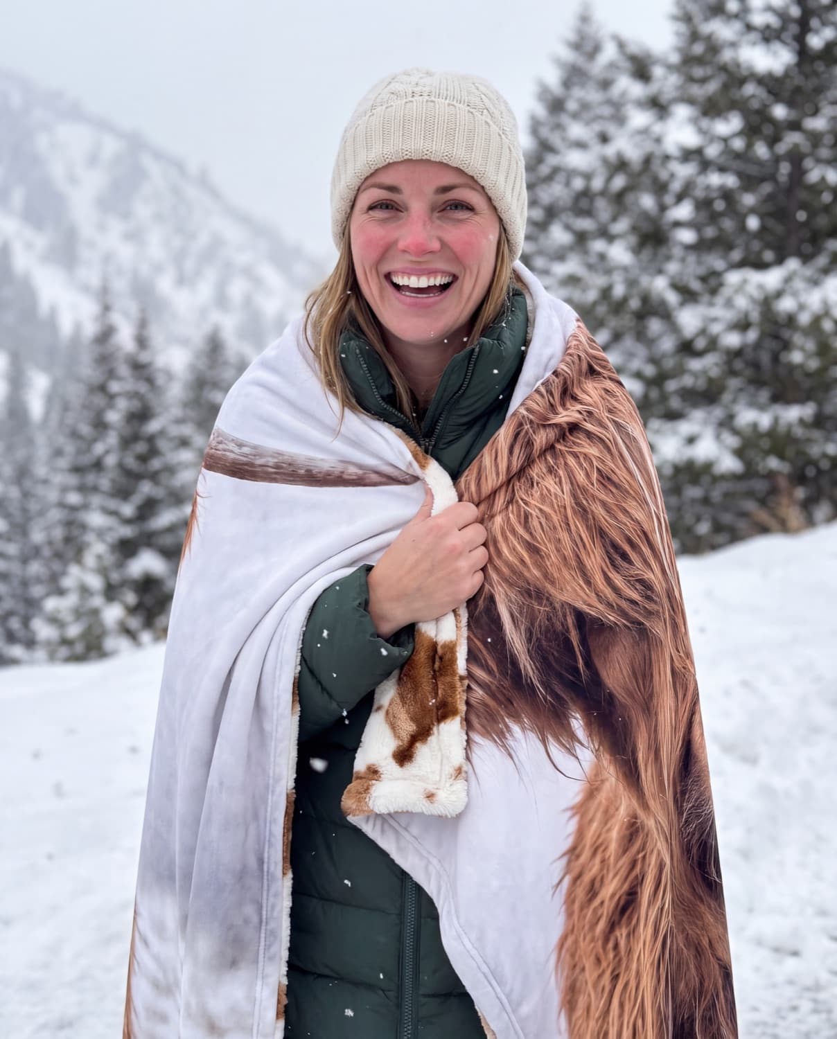 A laughing woman in a cream beanie at a snowy Utah canyon, wrapped in the Highland Snow blanket with its calf-patterned plush showing